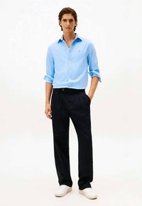 Giovane uomo in camicia azzurra chiara con bottoni, pantaloni neri, sneakers bianche, con una mano in tasca su uno sfondo bianco.