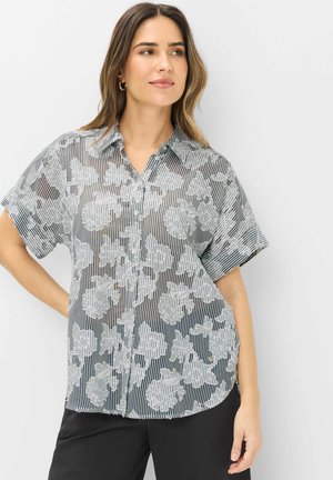 Donna con capelli lunghi che indossa una camicia trasparente con maniche corte e bottoni, con motivo floreale bianco e righe verticali, in piedi con la mano sul fianco.