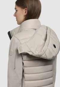 Lys beige jakke med en blød, puffet hætte og quiltet krop. Har en glat stofstruktur og et rundt logo-patch på ærmet.