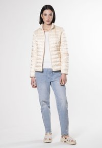 Giacca piumino beige chiaro con superficie texturizzata, zip centrale e colletto alto. Indossata sopra una maglietta bianca con jeans azzurri e sandali beige.
