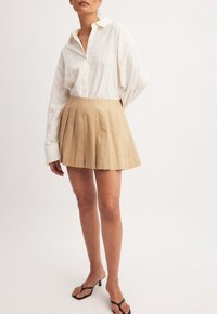 Chemise blanche à manches longues avec des rayures verticales, associée à une jupe mini plissée beige. Le mannequin porte des sandales à lanières noires.