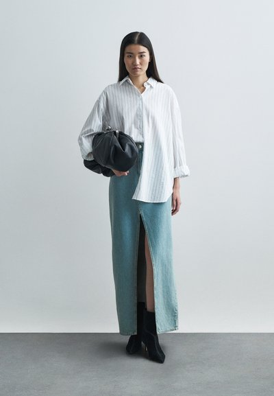 Cămașă albă cu dungi, cu mâneci largi, fustă maxi din denim deschis la culoare, cu un slit în față, și geantă mare, neagră. Botine negre.