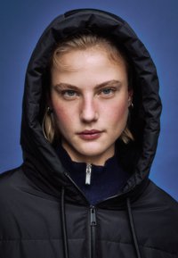 Musta hupullinen takki, jossa tikattu pinta ja vetoketjullinen suljenta, päällä tummansininen pooloneule. Mallilla on siniset silmät ja vaalea tukka.