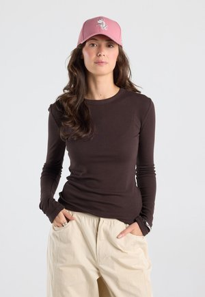 Vrouw met lang donker haar, een roze baseballpet, een nauwsluitend donker shirt met lange mouwen en lichtbeige broek, handen in de zakken.