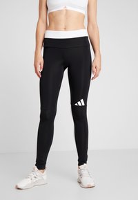 Czarne legginsy z wysokim stanem, wykonane z elastycznego materiału, z białym logo adidas na dolnej lewej nogawce.
