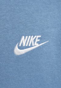 Vitt broderat Nike-logotyp med swoosh på en ljusblå stickad tygbakgrund.