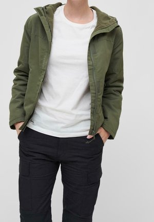 Personne portant une veste à capuche verte ouverte par-dessus un t-shirt blanc, les mains dans les poches d’un pantalon cargo noir.