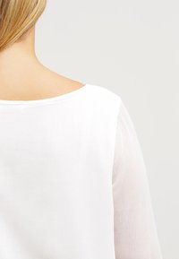Blusa blanca con escote amplio y mangas largas transparentes. La tela tiene una textura suave con finas líneas visibles, ofreciendo un aspecto ligero.