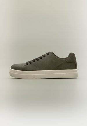 Baskets basses en daim gris foncé avec lacets noirs et semelle en caoutchouc blanche épaisse sur un fond clair neutre.