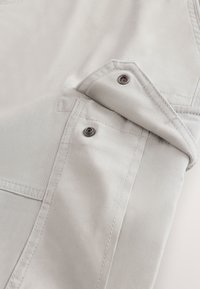 Tissu beige clair avec un rabat plié sécurisé par deux boutons-pression en métal et des détails visibles de surpiqûres sur un vêtement.