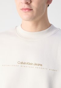 Krémová mikina s okrúhlym výstrihom vyrobená z mäkkej tkaniny. Obsahuje zlatý nápis: "Calvin Klein Jeans" a "ZALOŽENÉ V DEVäTNÁSTICH OSMDESIATYCH".