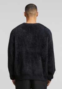 Suéter negro, peludo, con un diseño holgado, escote ancho y mangas largas; textura suave con un acabado de superficie ligeramente desigual.