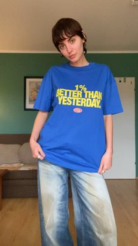 T-shirt azul oversized com texto amarelo a dizer "1% MELHOR DO QUE ONTEM." Mangas curtas, material de algodão, corte relaxado.