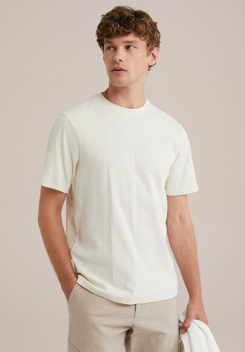 Baltas trumpas rankovėmis megztinis su tekstūruotais vertikaliais raštais ir apvaliu iškirpimu, derinamas su šviesiai smėlinėmis kelnėmis.