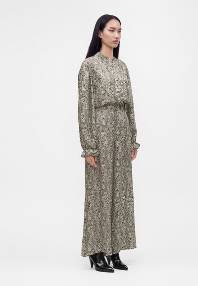 Femme debout portant une robe longue à manches longues, longueur cheville, avec un imprimé animal noir et beige et des bottines noires à talons.