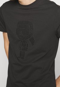 Camiseta de algodón negra con un gráfico brillante y texturizado de una figura con gafas de sol y una coleta, centrada en la parte delantera.