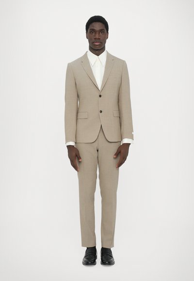 Beige op maat gemaakt pak met een enkelgeknoopt jasje, twee voorzakken en bijpassende broeken, gecombineerd met een witte overhemd met kraag en zwarte schoenen.
