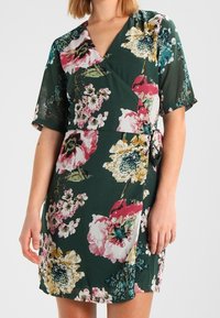Vestido envolvente floral con fondo verde, grandes flores rosas y amarillas, mangas cortas y un detalle de lazo en la cintura.