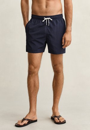 Man draagt marineblauwe zwemshorts met witte trekkkoord, staat op blote voeten in zwarte slippers tegen een neutrale achtergrond.