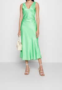 Satin grünes Kleid mit gewelltem Ausschnitt, tailliertem Oberteil und fließendem Saum. Kombiniert mit einer cremefarbenen Handtasche und Riemchenabsätzen.