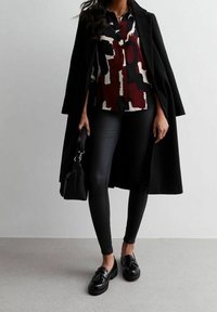 Donna che indossa un cappotto nero sulle spalle, blusa a fantasia, leggings neri, mocassini con nappine e tiene una borsa nera su sfondo grigio.
