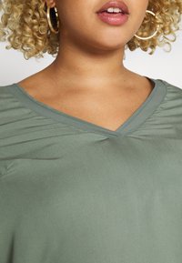 Top a V scollatura in tessuto verde tenue con delicati dettagli arricciati alle spalle. Texture liscia, senza motivi o accessori visibili.
