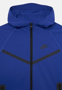 Chaqueta Nike azul con capucha, con un cierre negro y rayas de acento. El tejido parece suave con un ligero brillo.