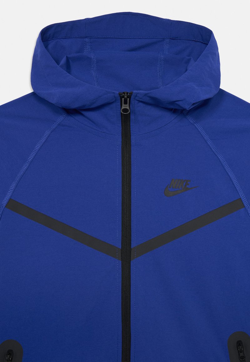 Chaqueta Nike azul con capucha, con un cierre negro y rayas de acento. El tejido parece suave con un ligero brillo.