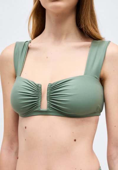 Ung kvinne iført grønn bikinitopp med brede stropper og en rynket sentral V-utskjæring på ensfarget bakgrunn.