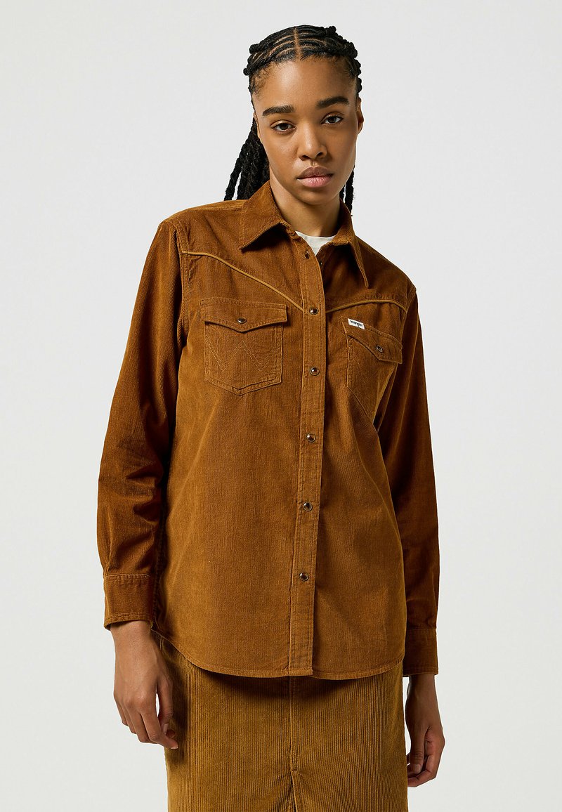 Chemise en velours côtelé marron à manches longues et col. Elle possède deux poches poitrine, des boutons-pression et une texture à côtes verticales.