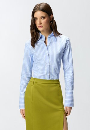 Donna con lunghi capelli castani che indossa una camicia a righe blu con bottoni e una gonna verde oliva con spacco laterale, in piedi davanti a uno sfondo semplice.