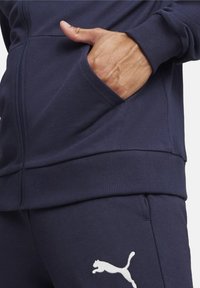 Felpa con zip blu navy e polsini a costine. Presenta una tasca frontale e un logo Puma bianco in basso a sinistra. Tessuto morbido e testurizzato.