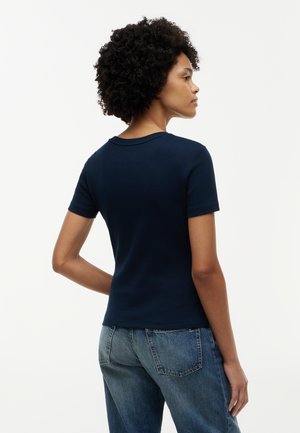 Kobieta o kręconych włosach, ubrana w ciemnoniebieską koszulkę z krótkim rękawem i niebieskie dżinsy, stoi tyłem na jednolitym jasnym tle.