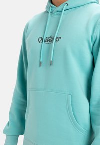 Sudadera azul claro con bolsillo frontal, cordón ajustable y logo de Quiksilver en impresión oscura en el pecho. Textura de material suave.