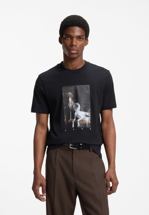 Ung mand iført en sort T-shirt med trykt billede af to hunde, kombineret med brune bukser og et sort bælte.