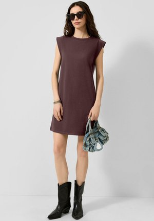 Frau trägt ärmelloses dunkelviolettes Kleid, schwarze Sonnenbrille, schwarze Stiefel und hält eine kleine blaue Denim-Handtasche vor einfarbigem Hintergrund.