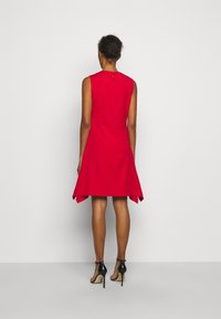 Röd ärmlös klänning med hög halsringning, utsvängd nederkant och detaljer med nyckelhål på ryggen, kombinerad med svarta högklackade skor.