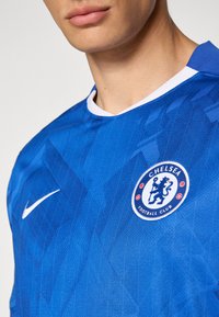 Blå Nike fodboldtrøje med struktureret geometrisk mønster, rund hvid krave og Chelsea Football Club-logo på venstre side.