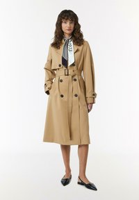 Karamelkleurige trenchcoat met een dubbele rij knopen, zwarte knopen en een ceintuur in de taille. Gecombineerd met een gestreepte sjaal en zwarte slingback schoenen.