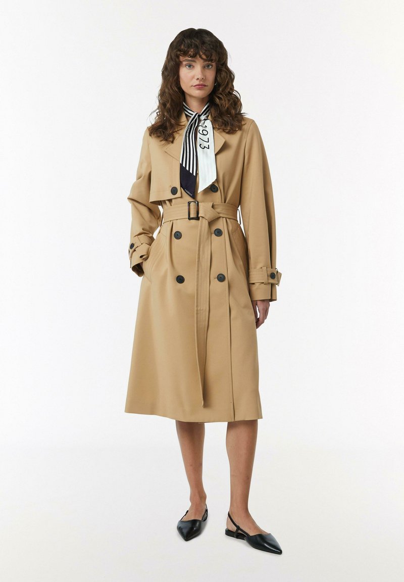 Karamelkleurige trenchcoat met een dubbele rij knopen, zwarte knopen en een ceintuur in de taille. Gecombineerd met een gestreepte sjaal en zwarte slingback schoenen.