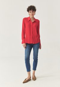 Camisa roja de manga larga con botones y cuello clásico, combinada con jeans azules ajustados y zapatos marrones de tipo slipper. Tejido suave, ajuste relajado.