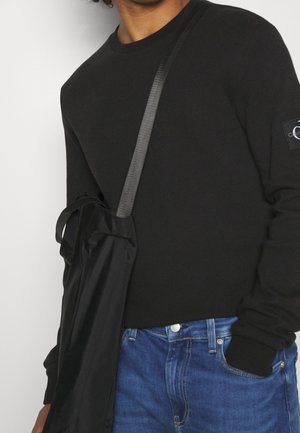 Personne portant un pull noir à manches longues et un jean bleu, avec un sac bandoulière noir à large sangle sur l'épaule.