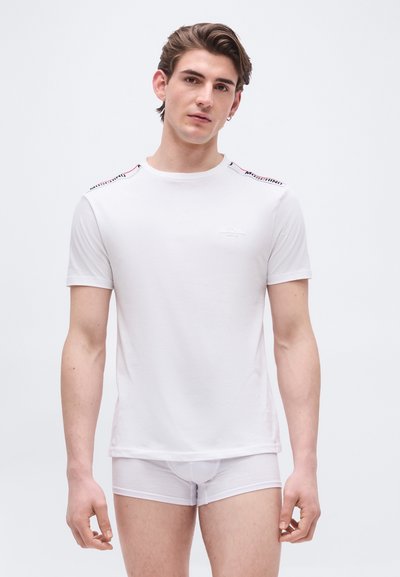 Jeune homme à la peau claire et aux cheveux bruns portant un t-shirt blanc Moschino et des boxer blancs assortis, debout devant un fond uni.