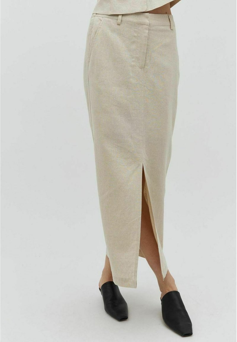 Jupe maxi en lin beige avec une fente frontale, dotée de poches latérales et d'une coupe ajustée. Associée à des mules noires à enfiler, mettant en valeur l'ensemble.