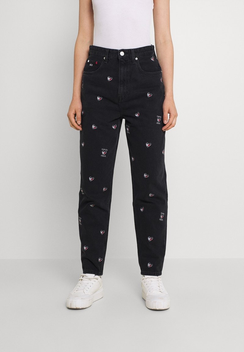 Vaqueros de mezclilla negros con patrones de corazones bordados en rojo y blanco, de talle alto, con corte recto, combinados con zapatillas blancas.