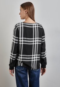 Sort og grå ternet bluse med lange ærmer, løs pasform og bådudskæring. Bæret sammen med denimjeans i blå, der har en subtil glans.