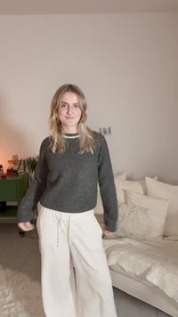 Maglione grigio lavorato a maglia con maniche lunghe e pantaloni larghi bianchi. Tessuto morbido con una vestibilità rilassata. Palette di colori neutri su sfondo di arredi morbidi.