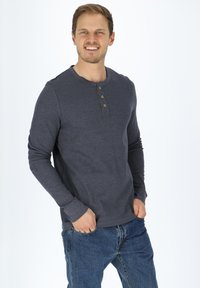 Mörkgrå långärmad T-shirt med henley-krage och tre bronsknappar. Bärs med blå jeans som har en avslappnad passform och mjuk textur.