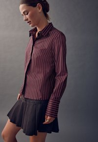 Bordó csíkos ing hosszú ujjú, gombos lezárással, fekete pliszírozott szoknyával párosítva, sima anyagú textúrával.