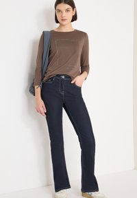 Brązowa koszula z długim rękawem z nadrukiem, w zestawieniu z ciemnoniebieskimi dzwonowatymi dżinsami z kontrastowym przeszyciem oraz casualowym plecakiem.